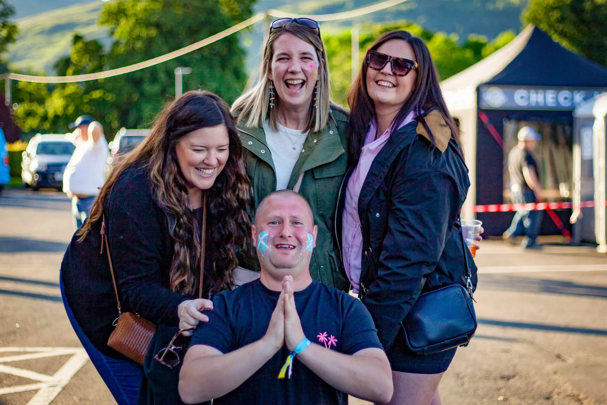 Four friends with saltire face paint laughing outside the festival check-in at golden hour